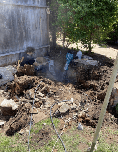 A person works on a large excavation in a backyard, using digging tools and surrounded by dirt and debris.